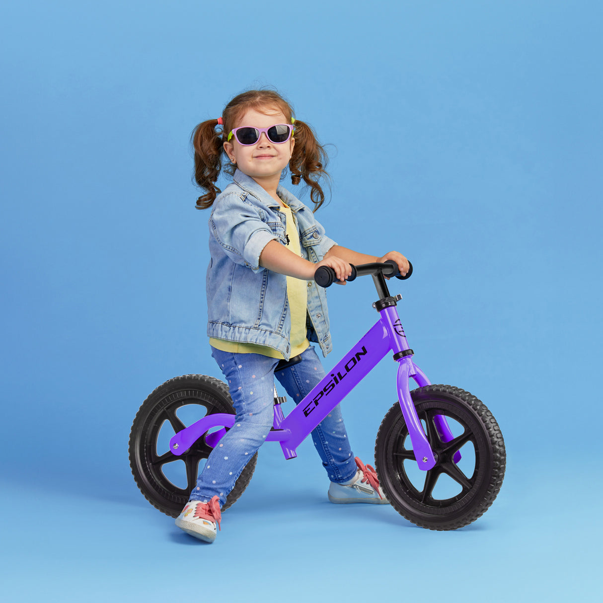 Young girl riding a purple Epsilon children's balance bike on blue background